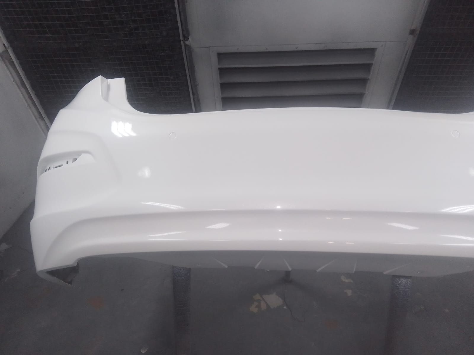 Parachoques de coche de color blanco, recién pintado en taller, colocado debajo de un sistema de ventilación.