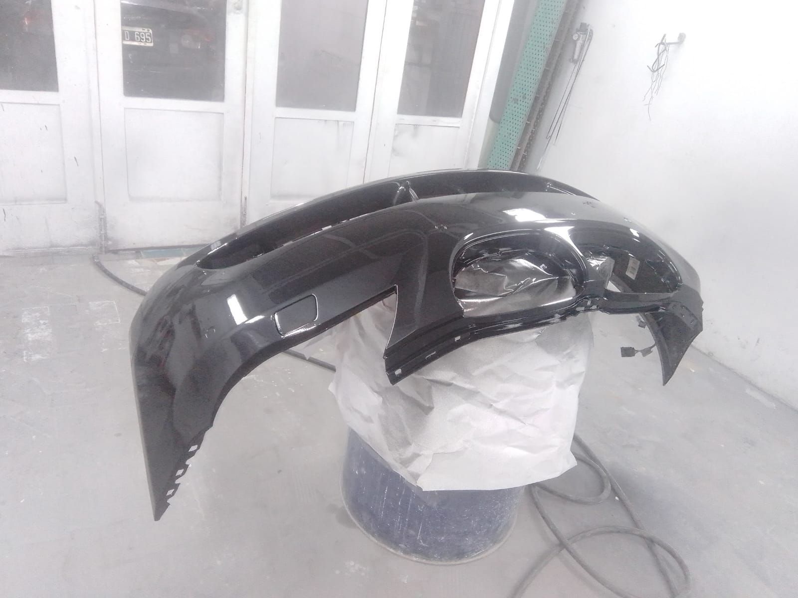 Parachoques de coche negro, dañado, apoyado sobre una bolsa blanca sobre un cubo azul, en un taller.