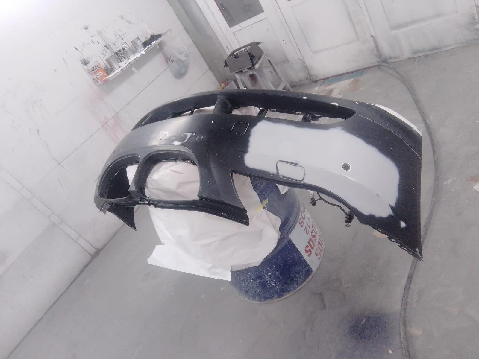 Parachoques de coche, parcialmente pintado de negro, apoyado sobre un cubo azul en el interior de un taller.