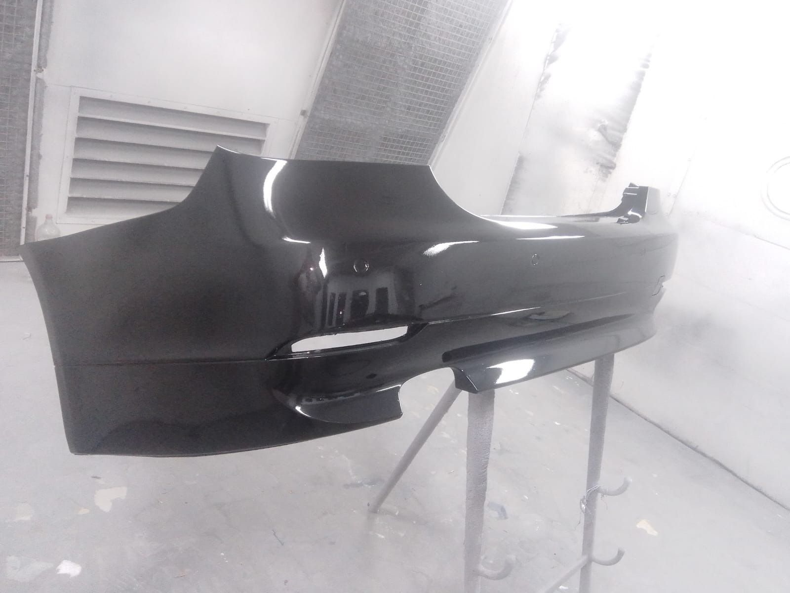 Parachoques de coche negro recién pintado en cabina de pintura, apoyado sobre un soporte.