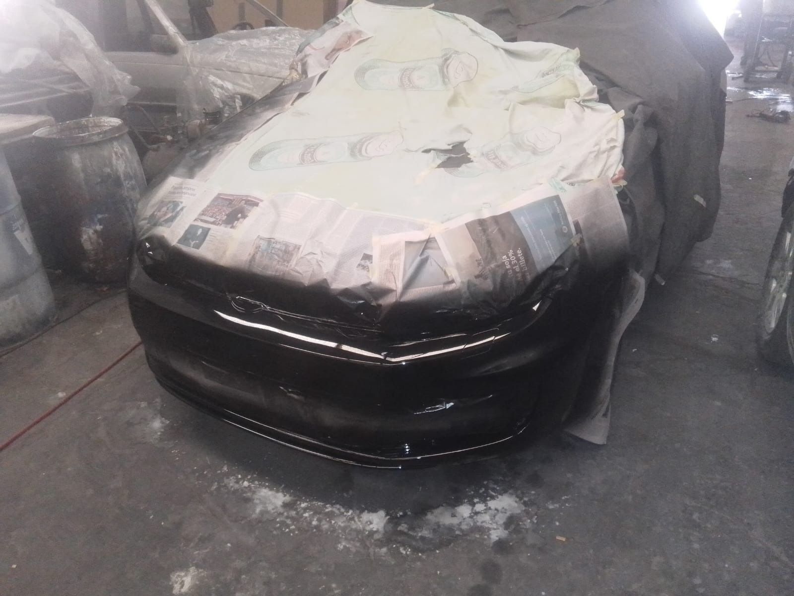 Parachoques de coche negro siendo pintado en un taller; áreas cubiertas y enmascaradas con periódico y papel.