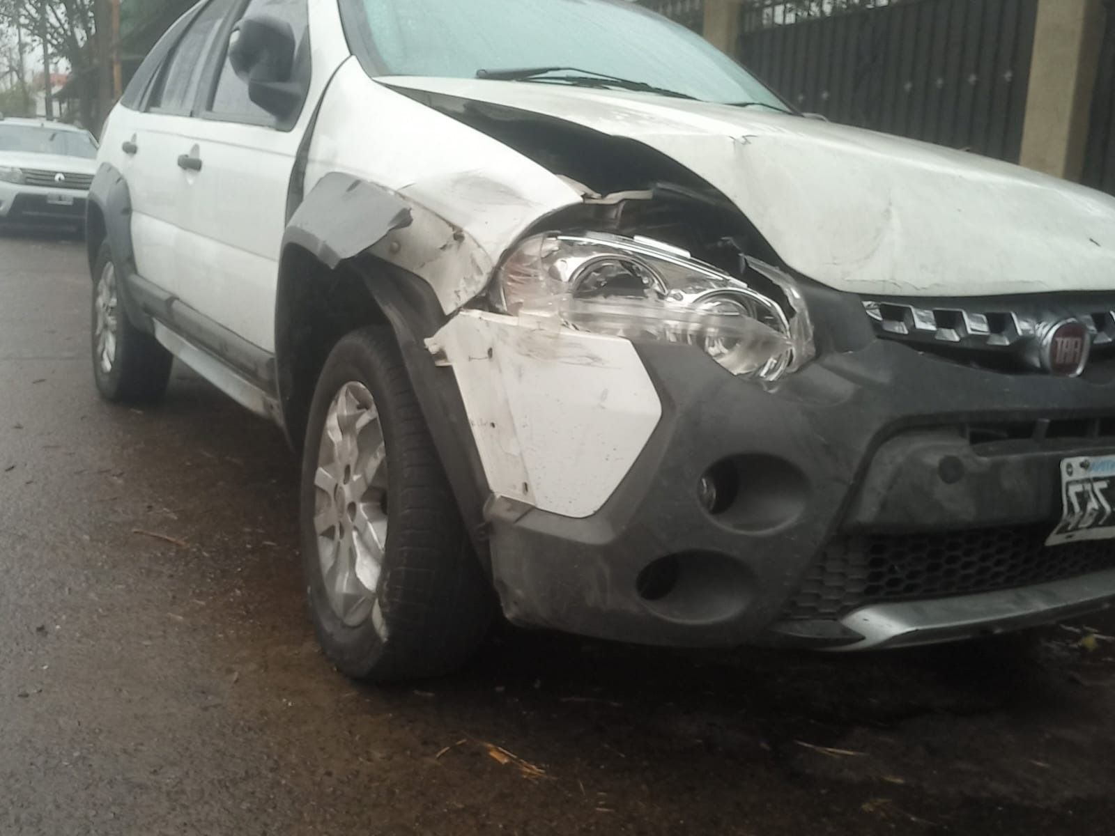 Automóvil blanco con daños en el frontal, capó abollado y faro roto, estacionado en una calle.