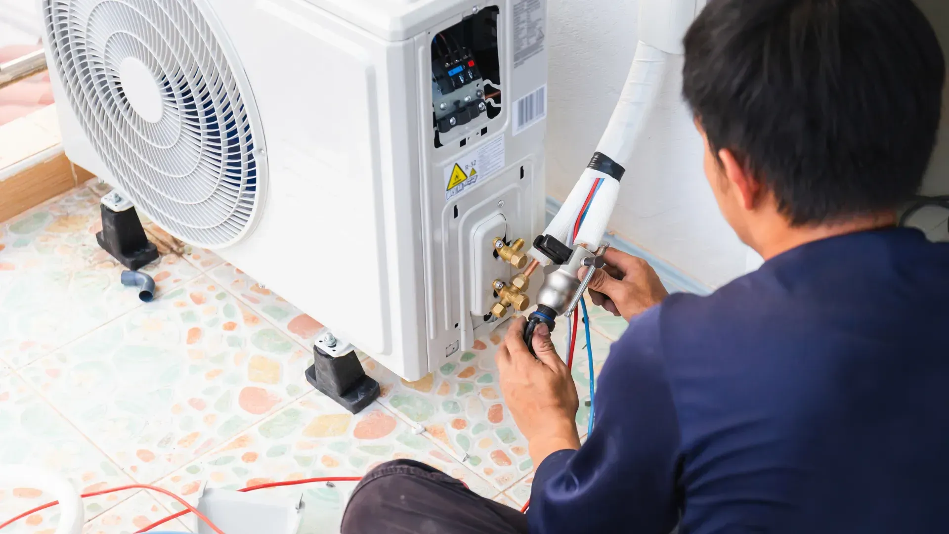 Man installing an AC unit outdoors, kneeling and working on the pipes near the condenser.