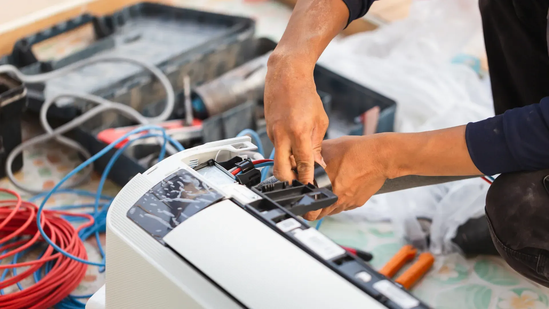 Person repairs an air conditioner, surrounded by tools and wires.