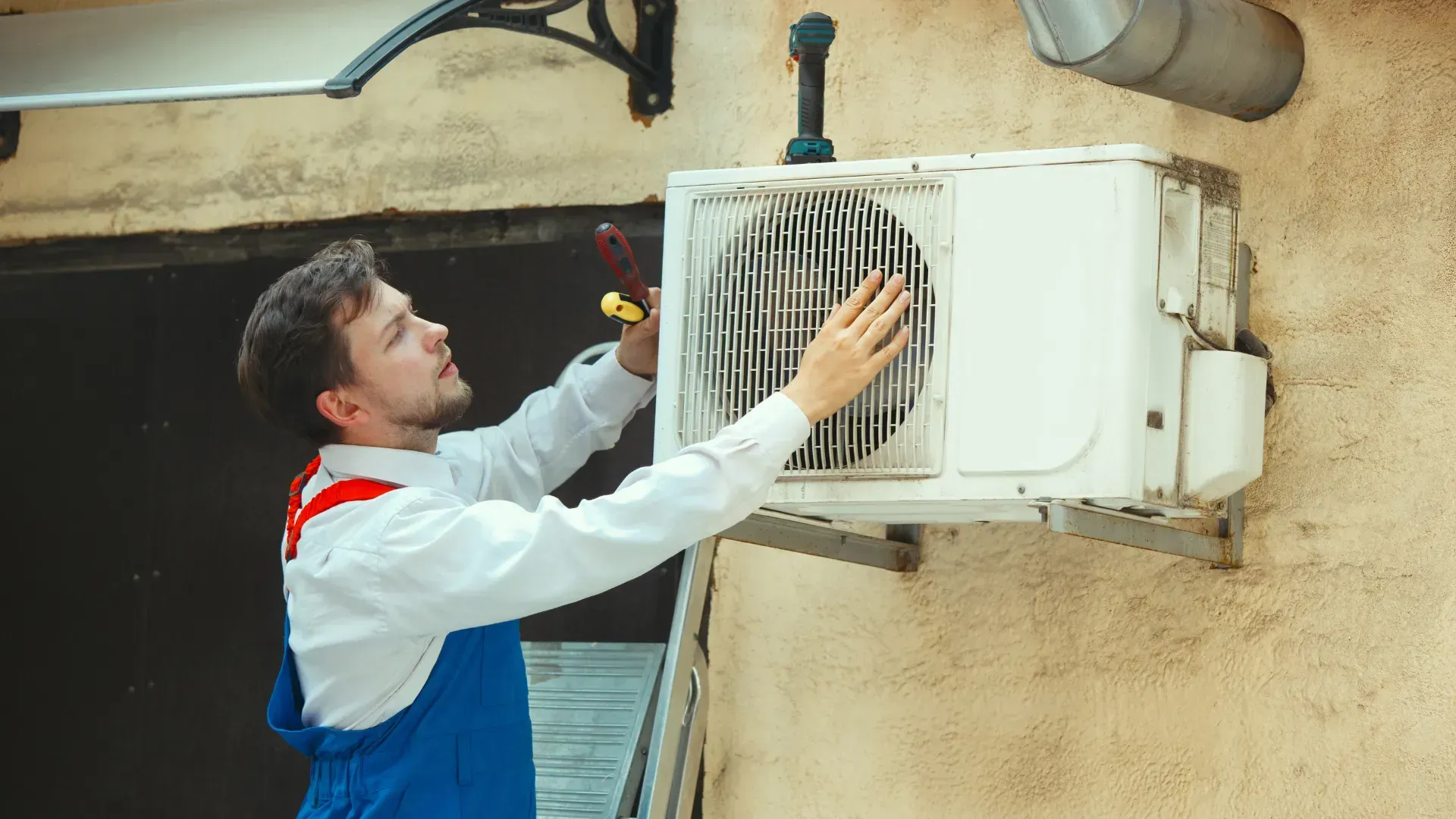 A technician in blue overalls repairs an outdoor air conditioning unit on a building's exterior.