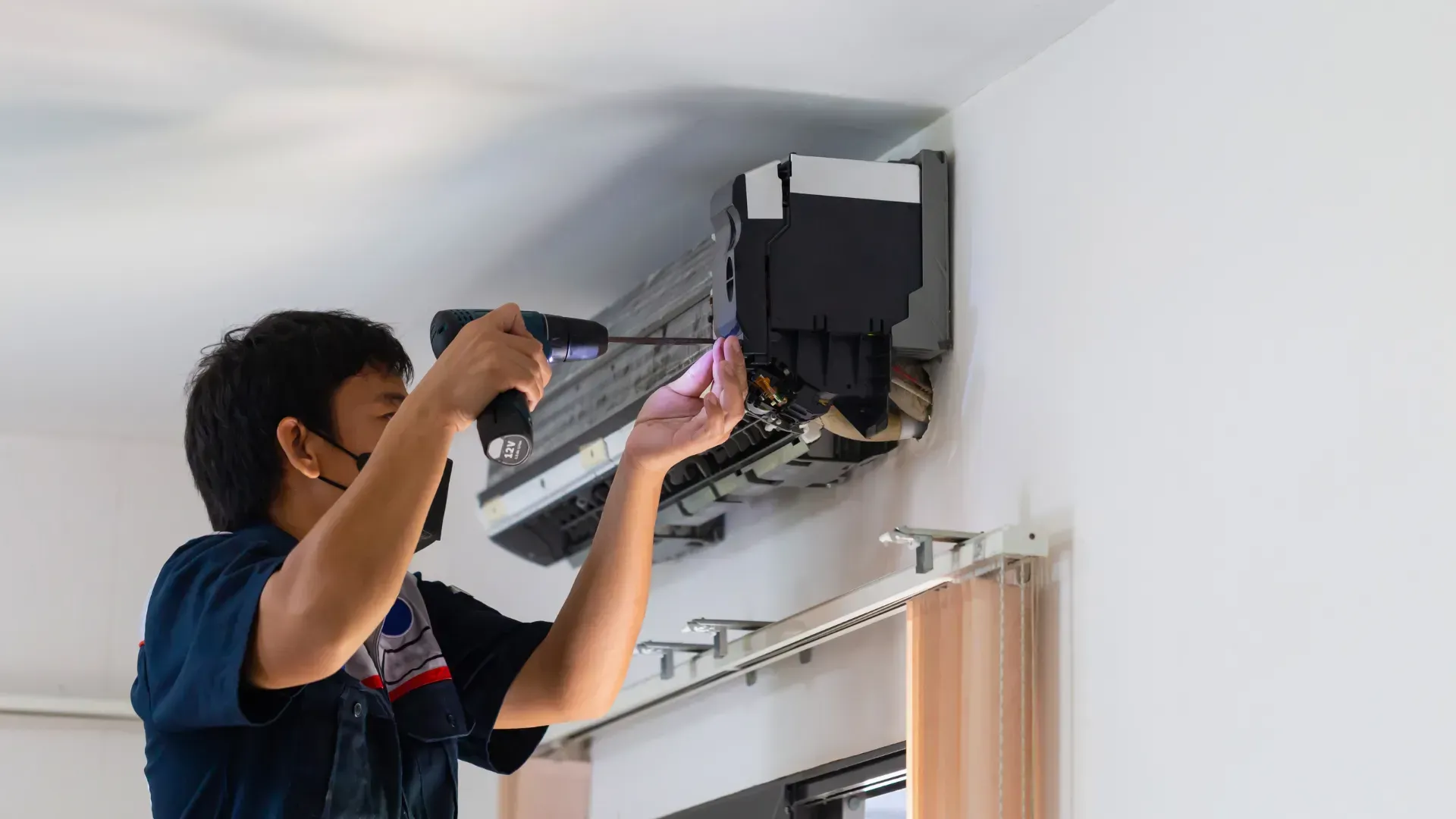 Person wearing a mask fixing an AC unit on a white wall, using a power drill.