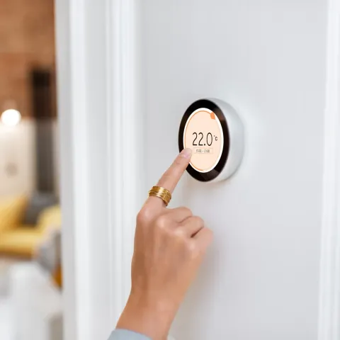 A hand touching a smart thermostat set to 22.0 degrees Celsius on a white wall.