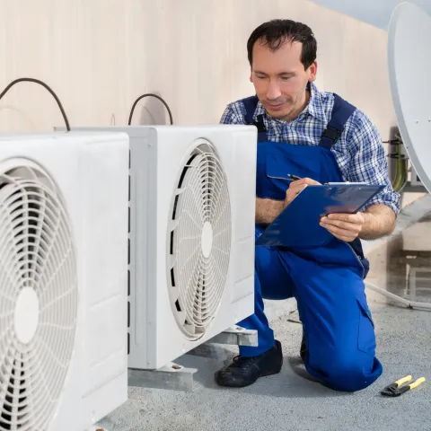 HVAC technician in blue overalls, inspecting air conditioning unit, writing on a clipboard outside.