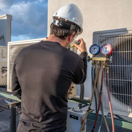 HVAC technician in white hard hat repairs an outdoor AC unit on a rooftop, using gauges.