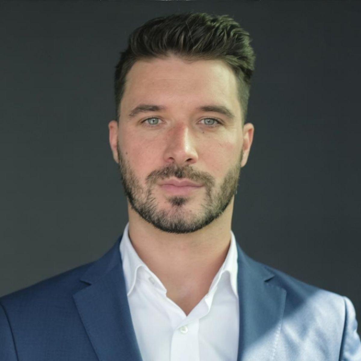 Man in blue suit and white shirt, close-up portrait, looking forward, short dark hair, stubble.