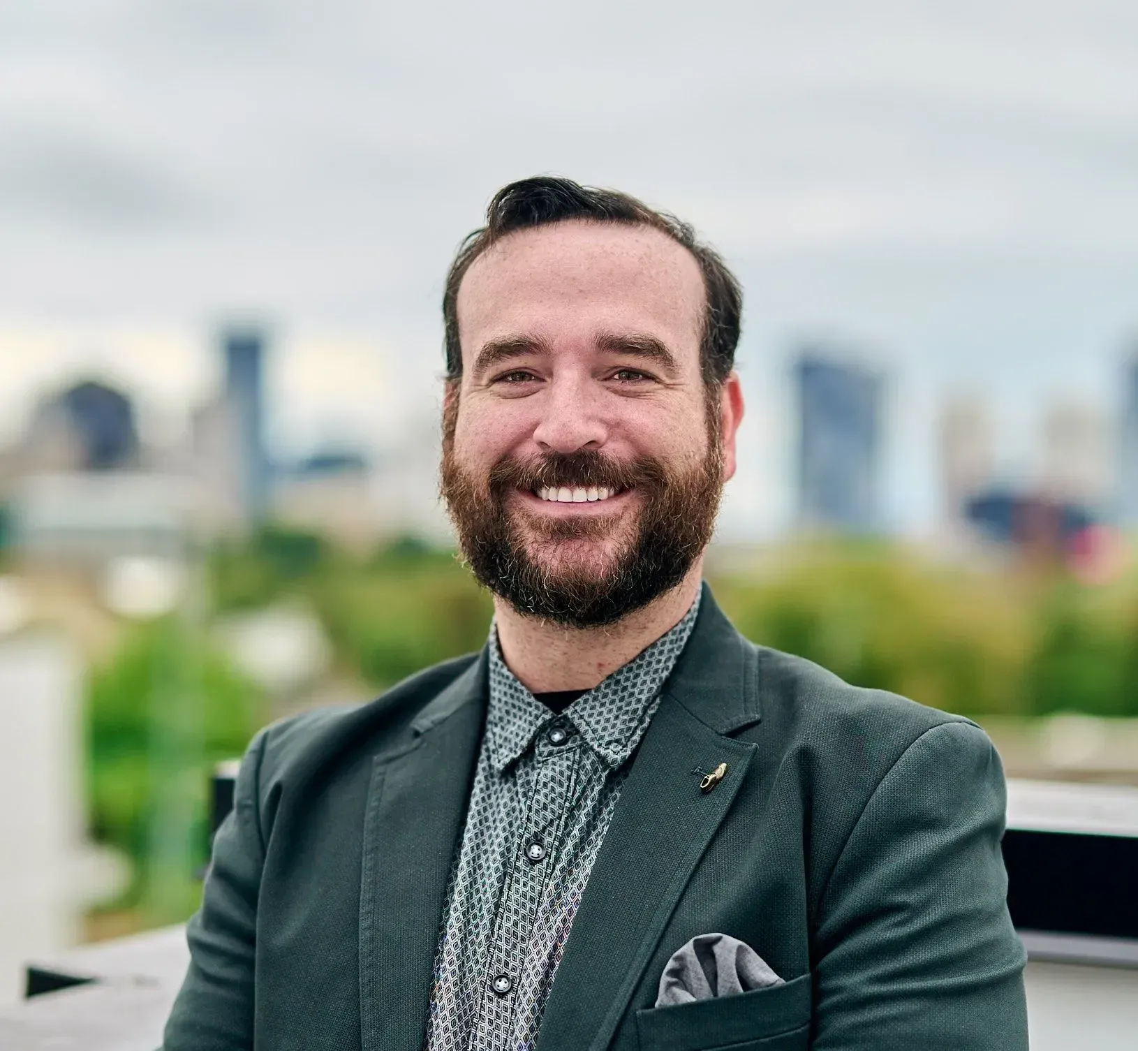 Man with a beard in a green blazer smiles outdoors with city backdrop.