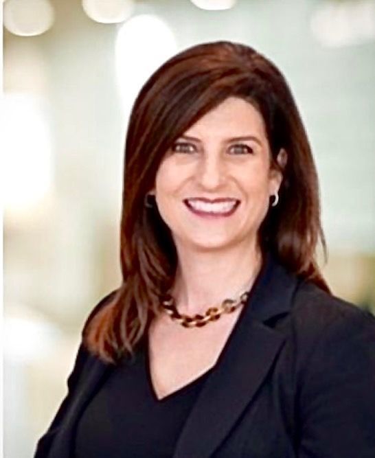 Woman with dark hair smiles, wearing a black blazer and necklace, against a blurred background.