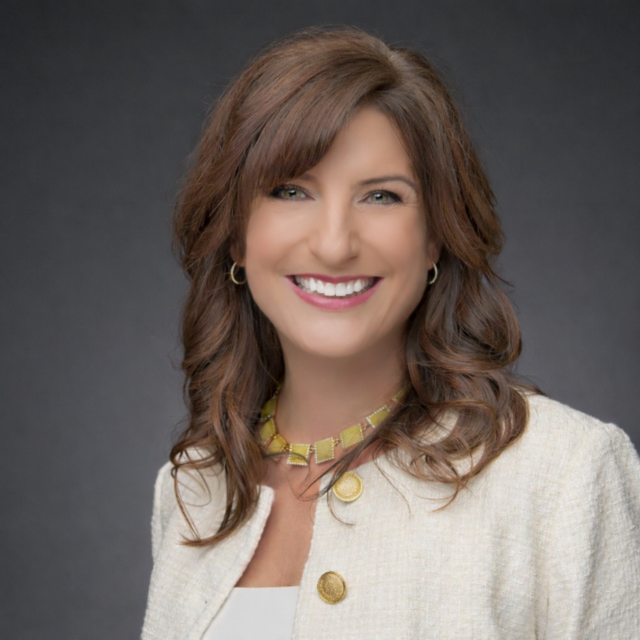 Woman with brown hair smiles, wearing a black blazer and necklace, against a blurry background.