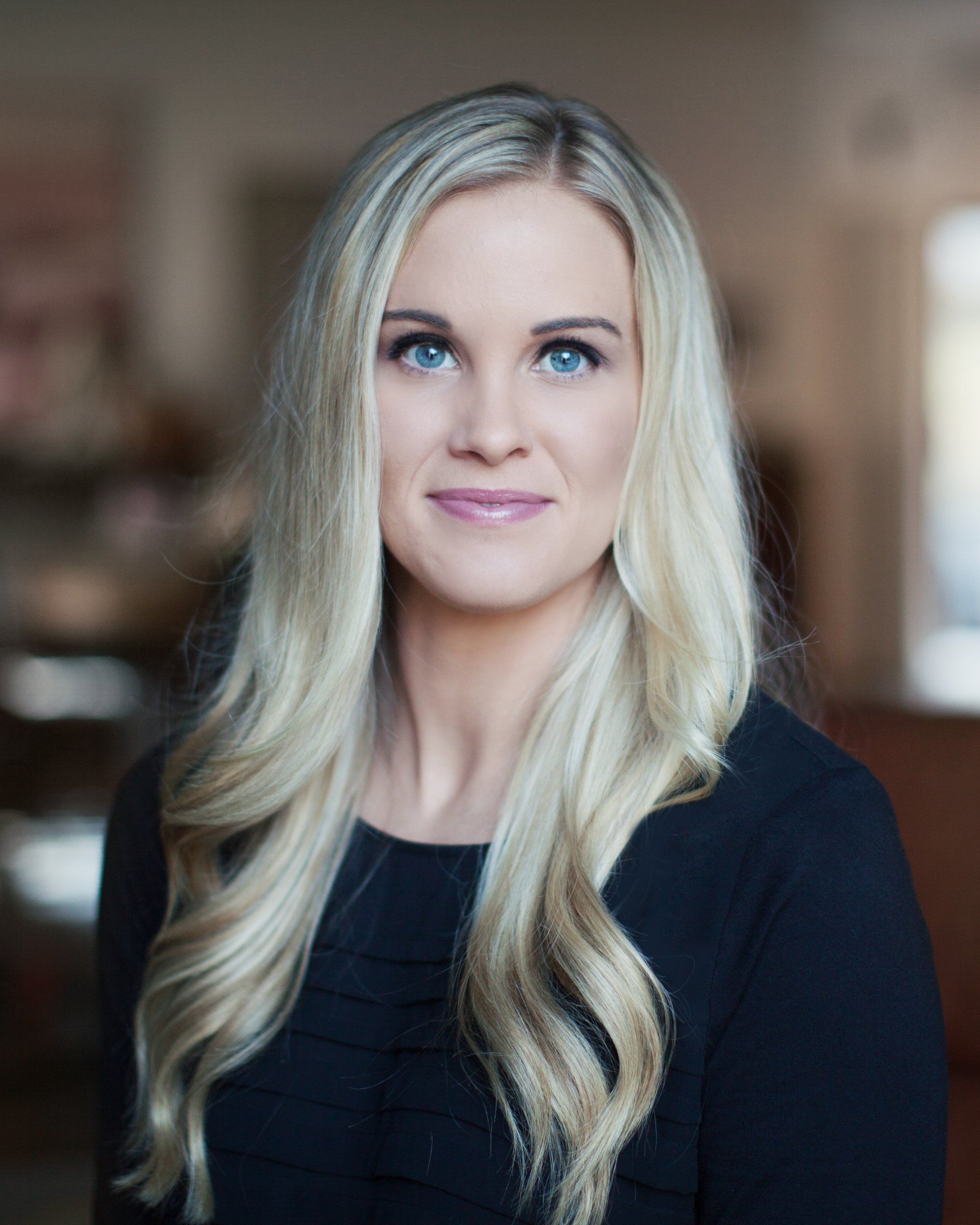 Blonde woman with blue eyes smiles, wearing a black top, indoors.