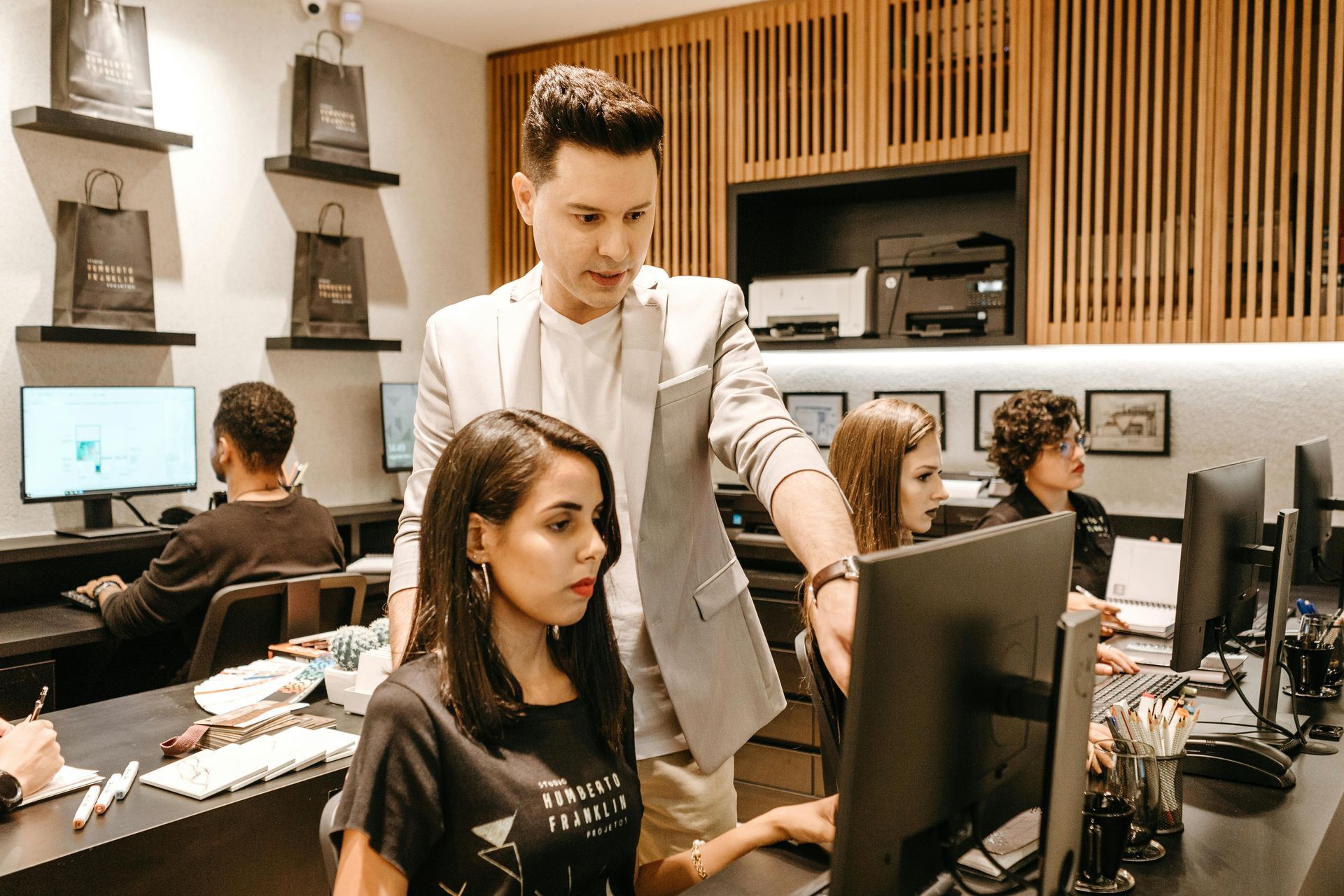 Man pointing at computer screen, instructing woman in a modern office setting, other workers at desks.