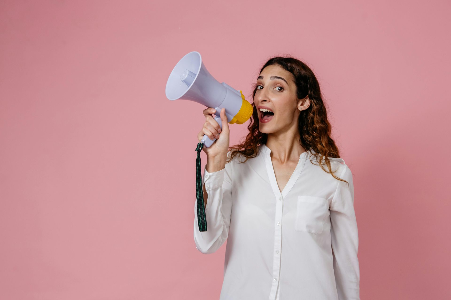 Woman in white shirt with curly hair, shouting into a megaphone against a pink background.
