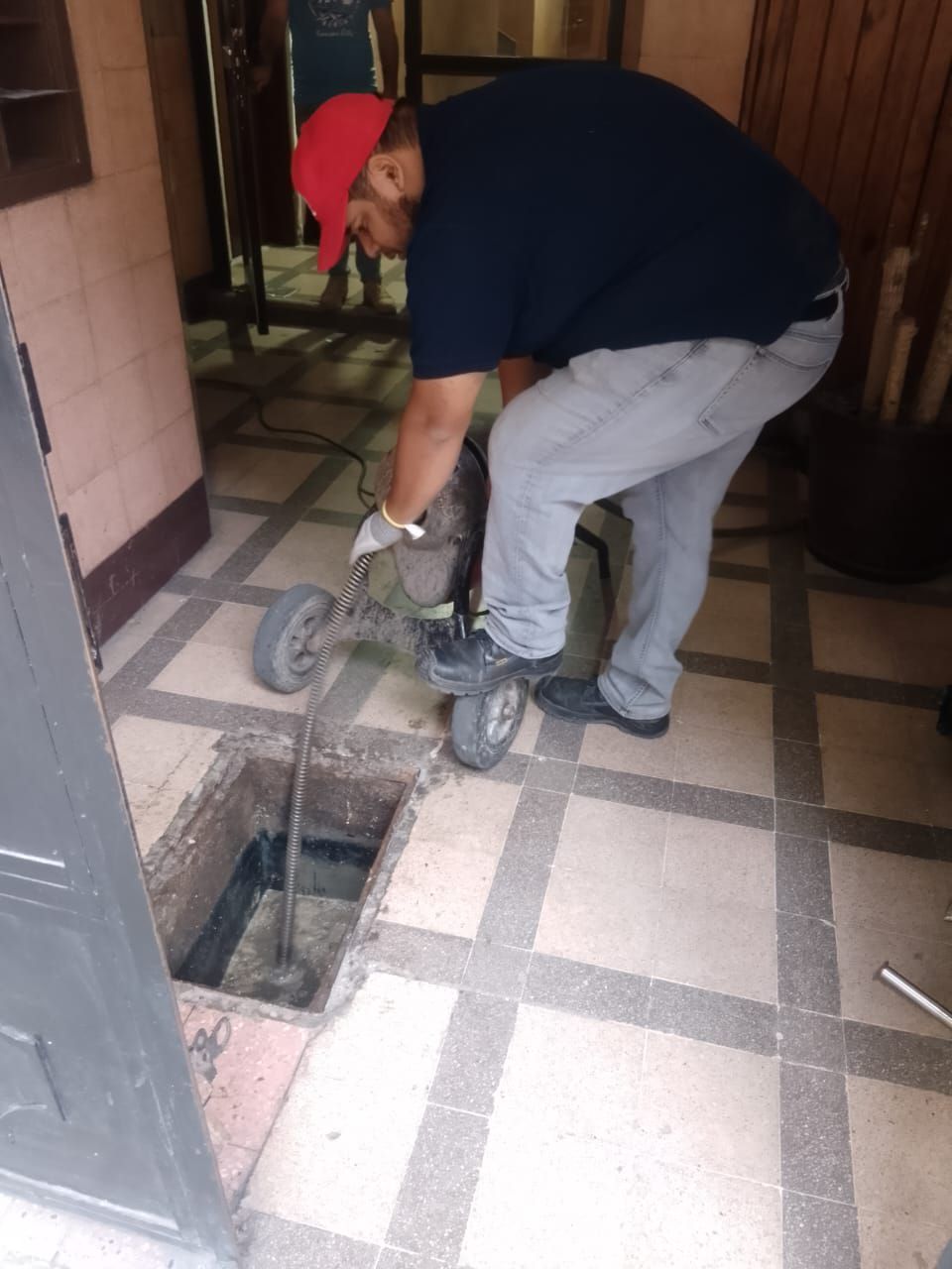 Hombre utilizando una máquina desatascadora cerca de una alcantarilla abierta. Interior con azulejos estampados. Gorra roja, camisa azul.
