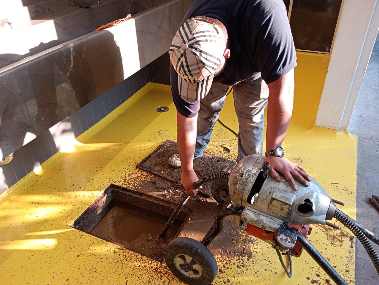 Hombre utilizando una máquina desatascadora, de pie en una zona pintada de amarillo, con un desagüe abierto a la vista.