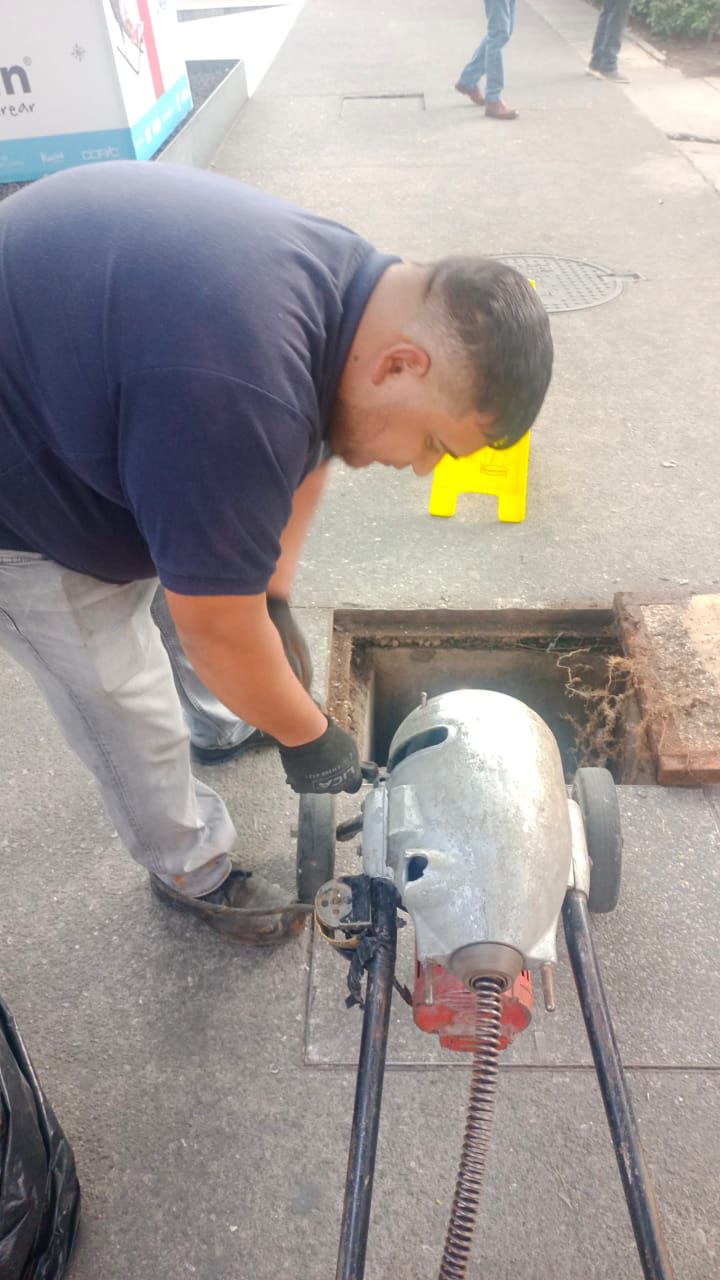 Hombre con camisa azul, utilizando una máquina desatascadora cerca de una alcantarilla callejera.