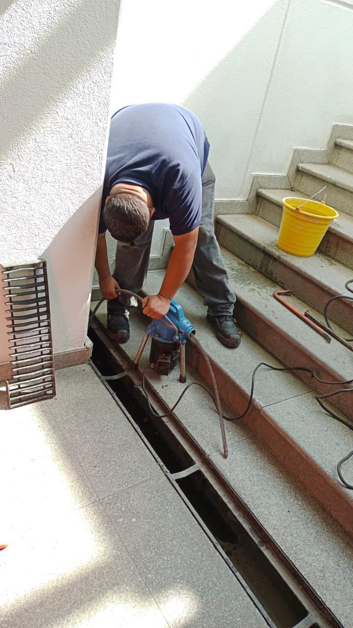Hombre utilizando una herramienta eléctrica para trabajar en una rejilla metálica en unos escalones cerca de una pared; cubo amarillo presente.