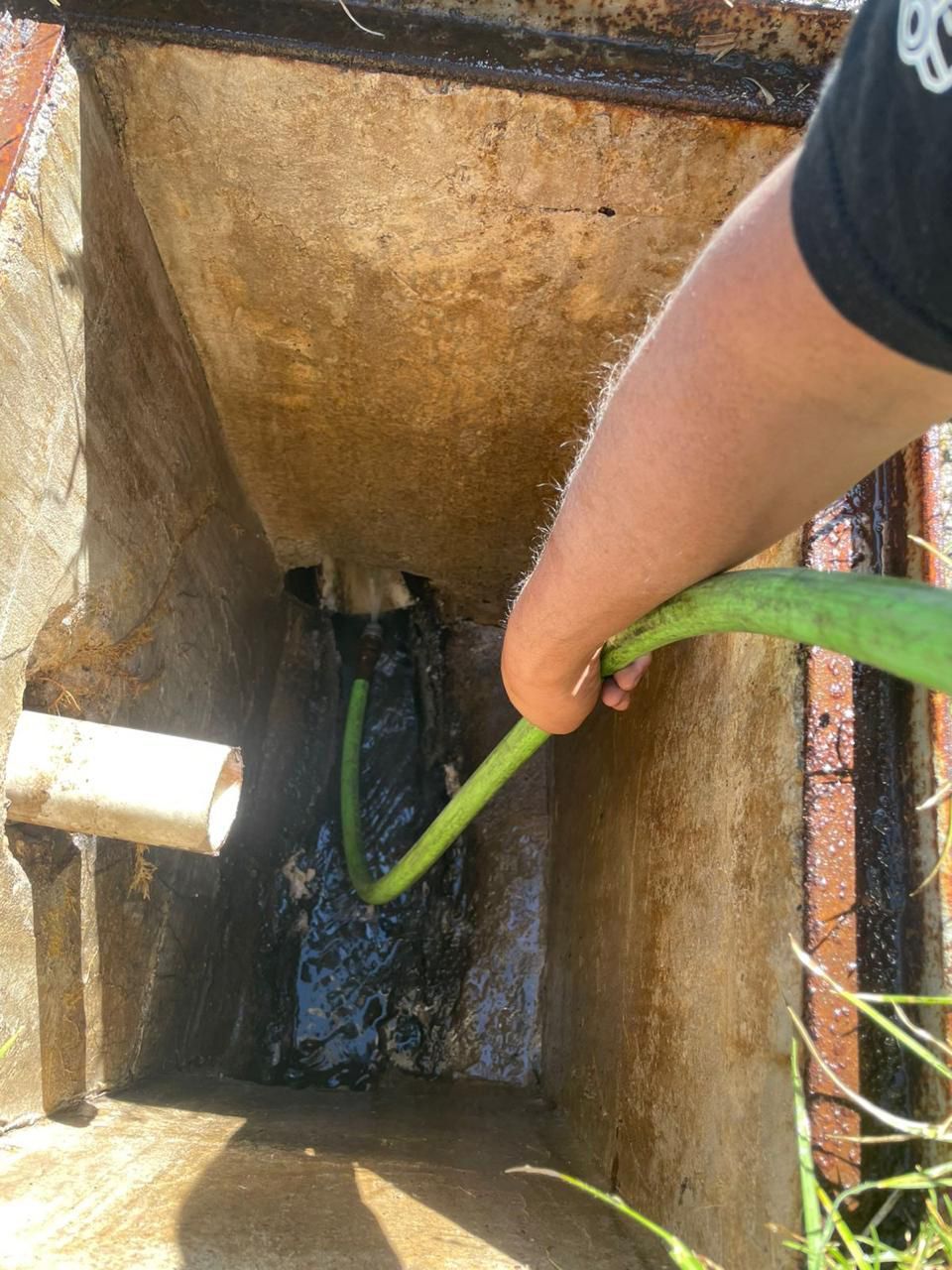 Una persona utiliza una manguera verde para eliminar los residuos de un desagüe de hormigón en un entorno exterior.