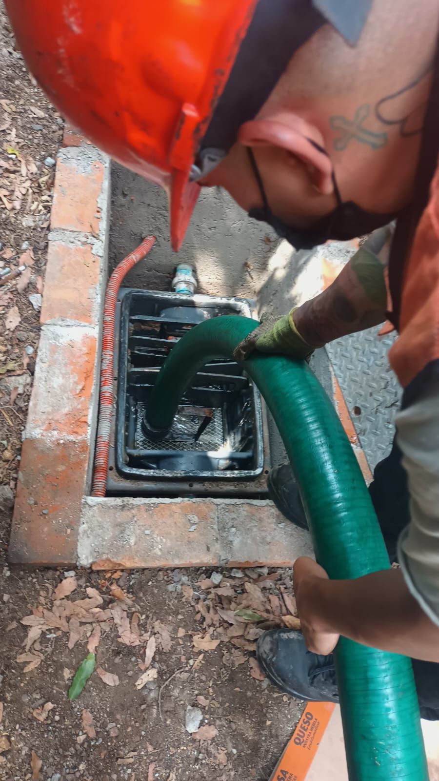 Hombre con casco de seguridad introduce una manguera verde en un desagüe oscuro dentro de un recinto de ladrillo.