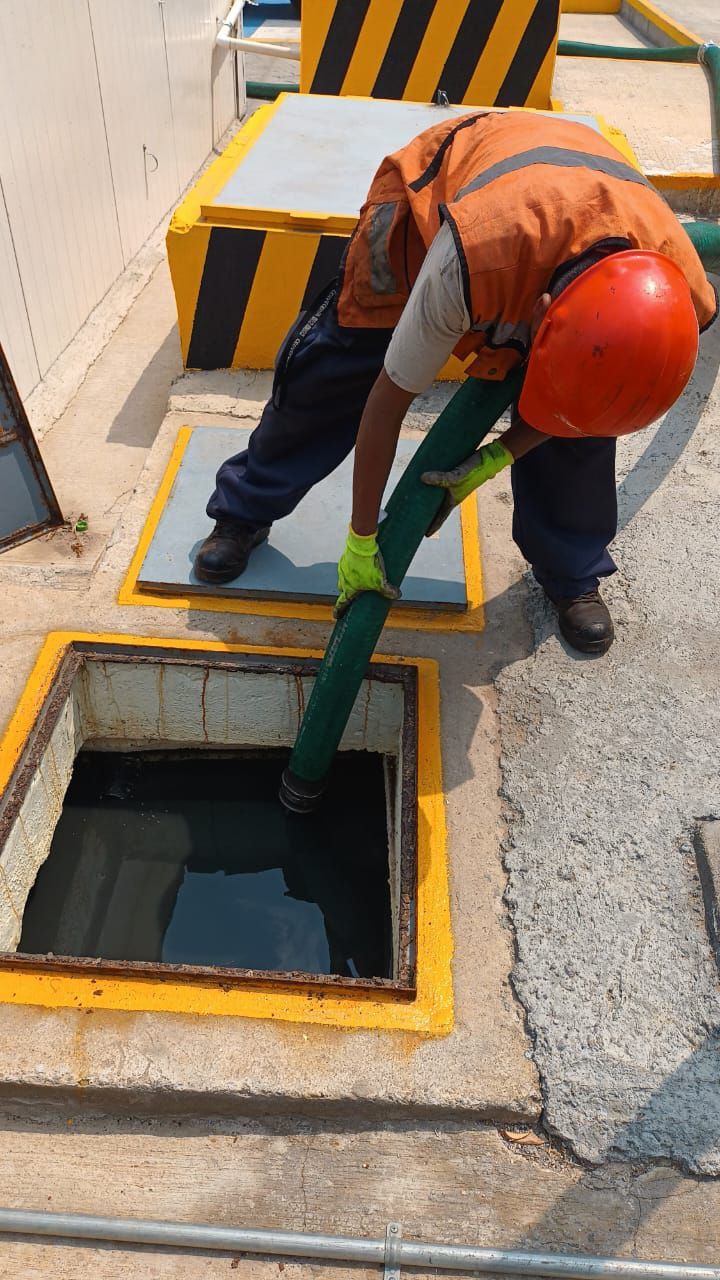 Trabajador con chaleco y casco naranjas sosteniendo una manguera verde, extrayendo líquido de un tanque abierto.
