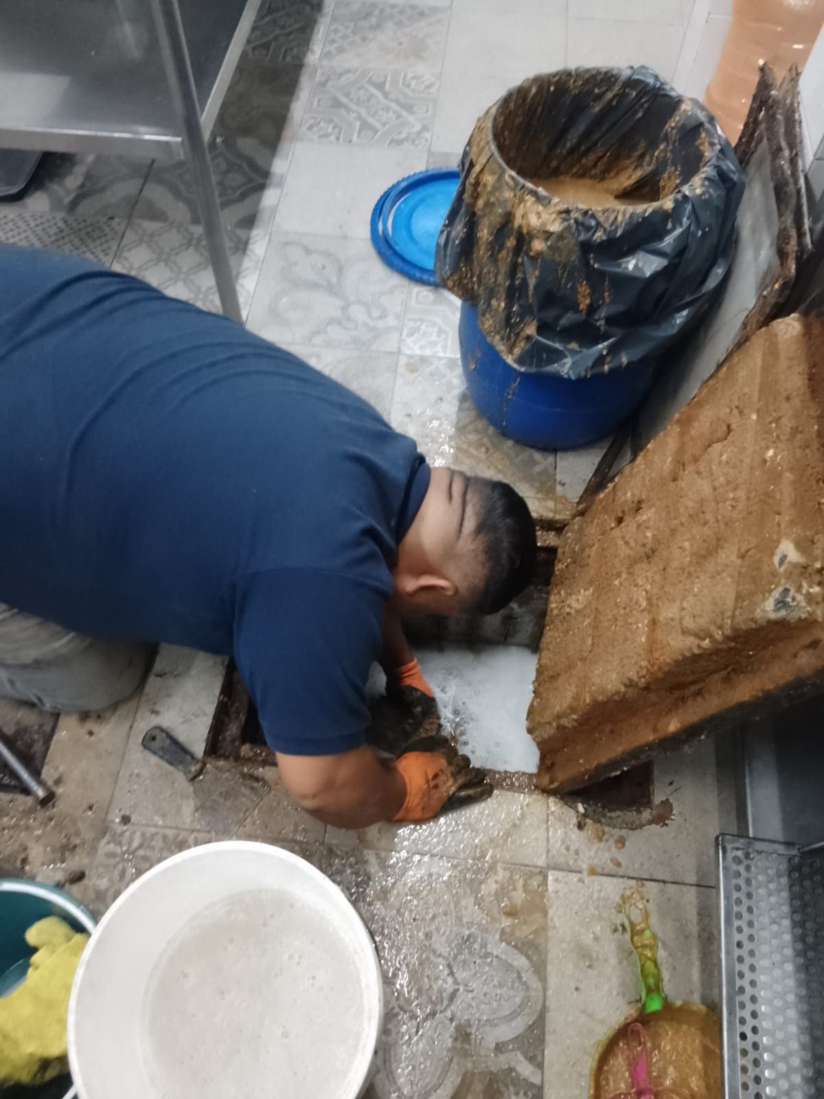 Hombre con camisa azul trabaja en un desagüe del suelo con un cubo y escombros cerca. Entorno de cocina.