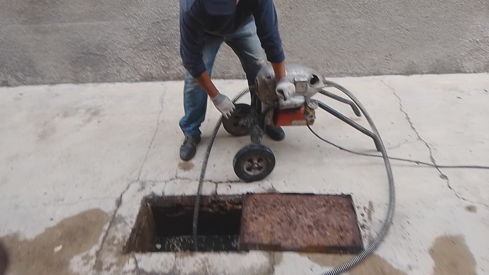 Una persona utiliza una máquina desatascadora para destapar una alcantarilla. El equipo se encuentra junto a una tapa de alcantarilla abierta.