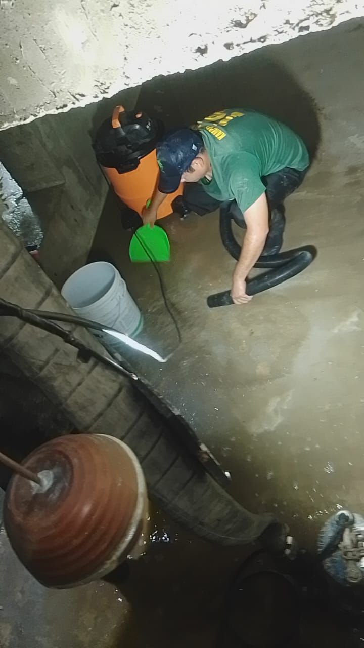 Una persona con camisa verde aspira el agua de un sótano inundado. Cerca hay una aspiradora industrial naranja y un cubo.