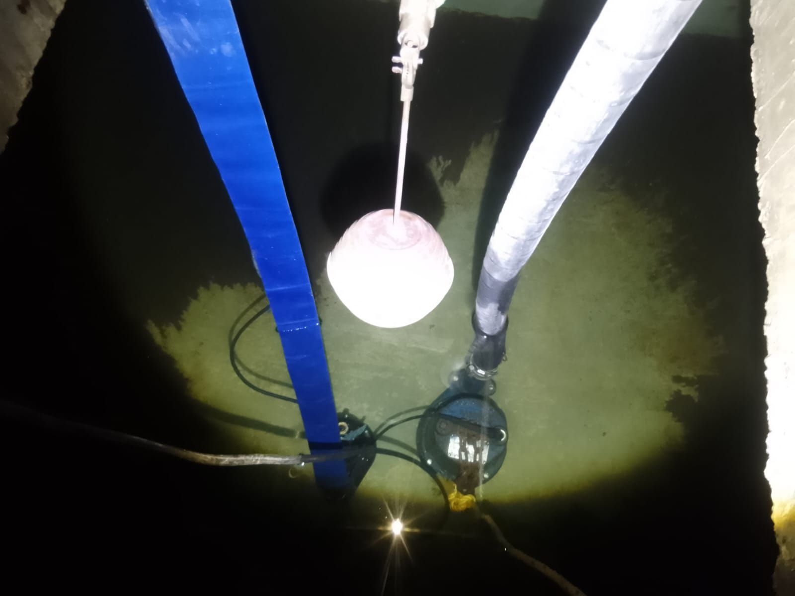 Dentro de un tanque oscuro, un flotador blanco cuelga sobre una bomba sumergida. Una correa azul y una manguera gris salen de la parte superior.