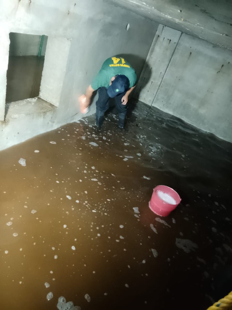 Una persona con camisa y gorra verdes limpia una zona de hormigón inundada. Un cubo rosa está cerca.