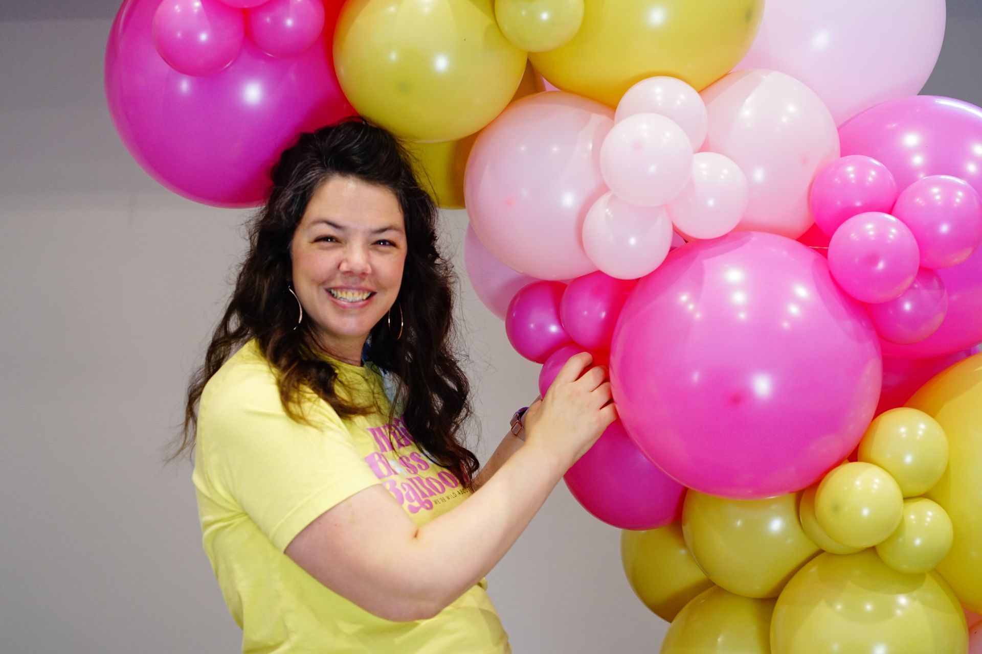 A woman in a yellow shirt is decorating a bunch of pink and yellow balloons.