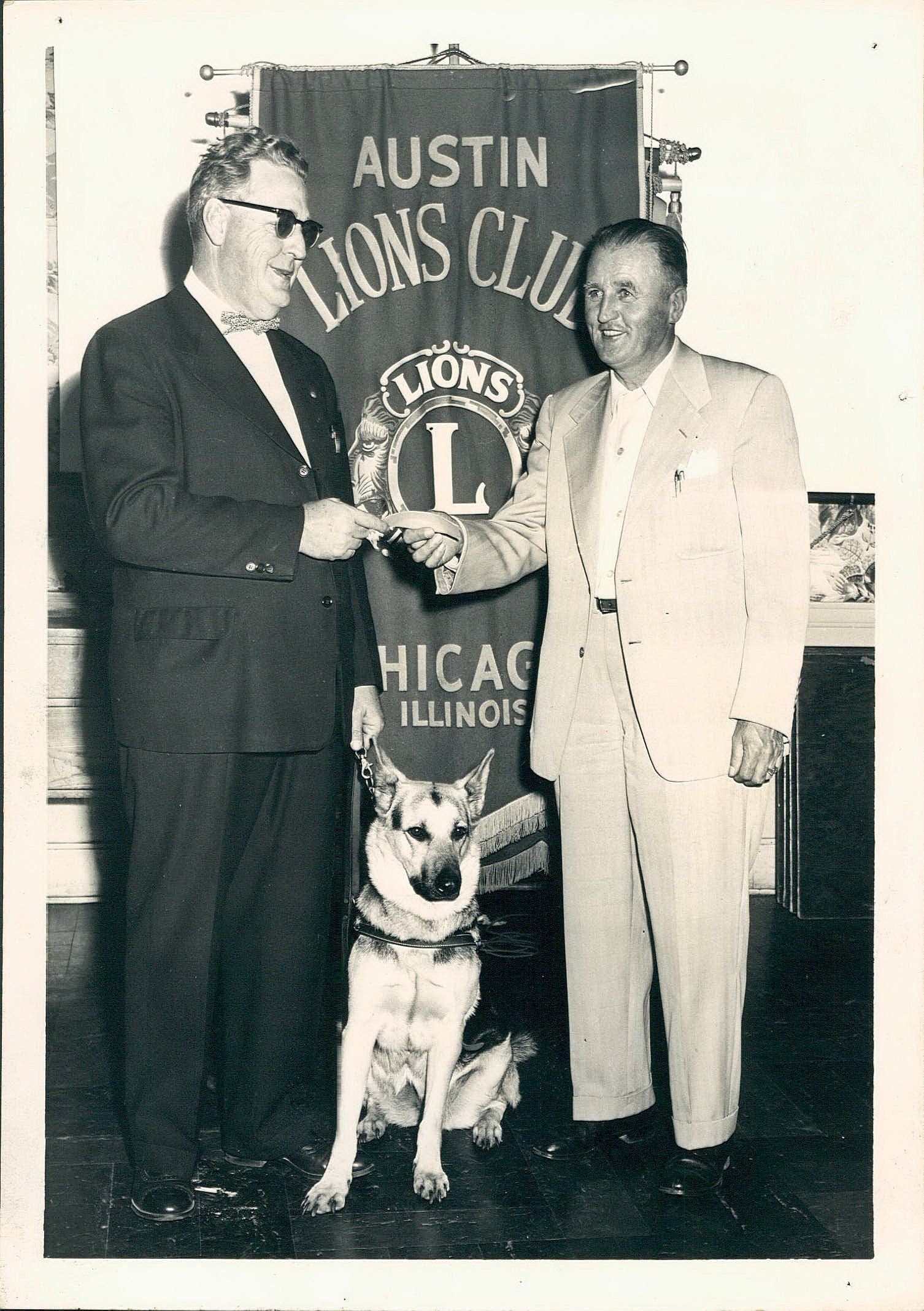 Two men shaking hands with a dog in front of a banner that says austin lions club