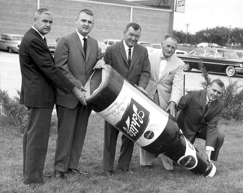 A group of men in suits are standing around a large bottle of soda