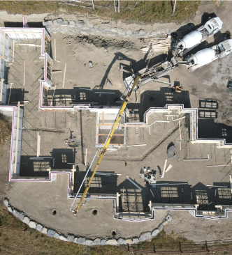 Aerial view of a residential foundation under construction with a long yellow concrete pump boom extending over the site.