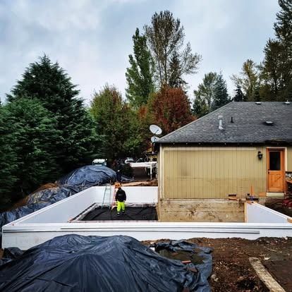 A construction worker stands inside a rectangular white ICF form foundation next to a house under renovation.