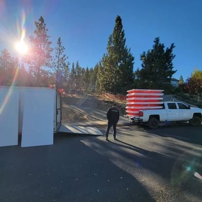 A person stands between a parked white truck loaded with stacked orange barriers and a utility trailer on a sunny day.