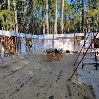 Construction site with white foam wall panels held up by metal supports on a wooden floor in a forest setting.