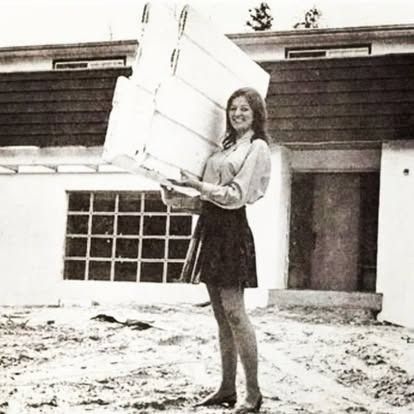 A smiling person standing on a dirt lot in front of a house, holding a large stack of white rectangular building panels.