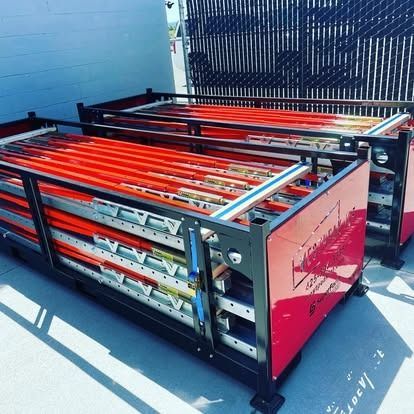 Two stacked metal shipping crates containing multiple orange-painted scaffolding or ladder components on an outdoor lot.
