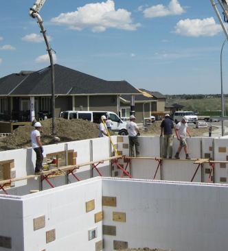 Construction workers in hard hats stand on scaffolding above white foam forms for a residential concrete foundation.