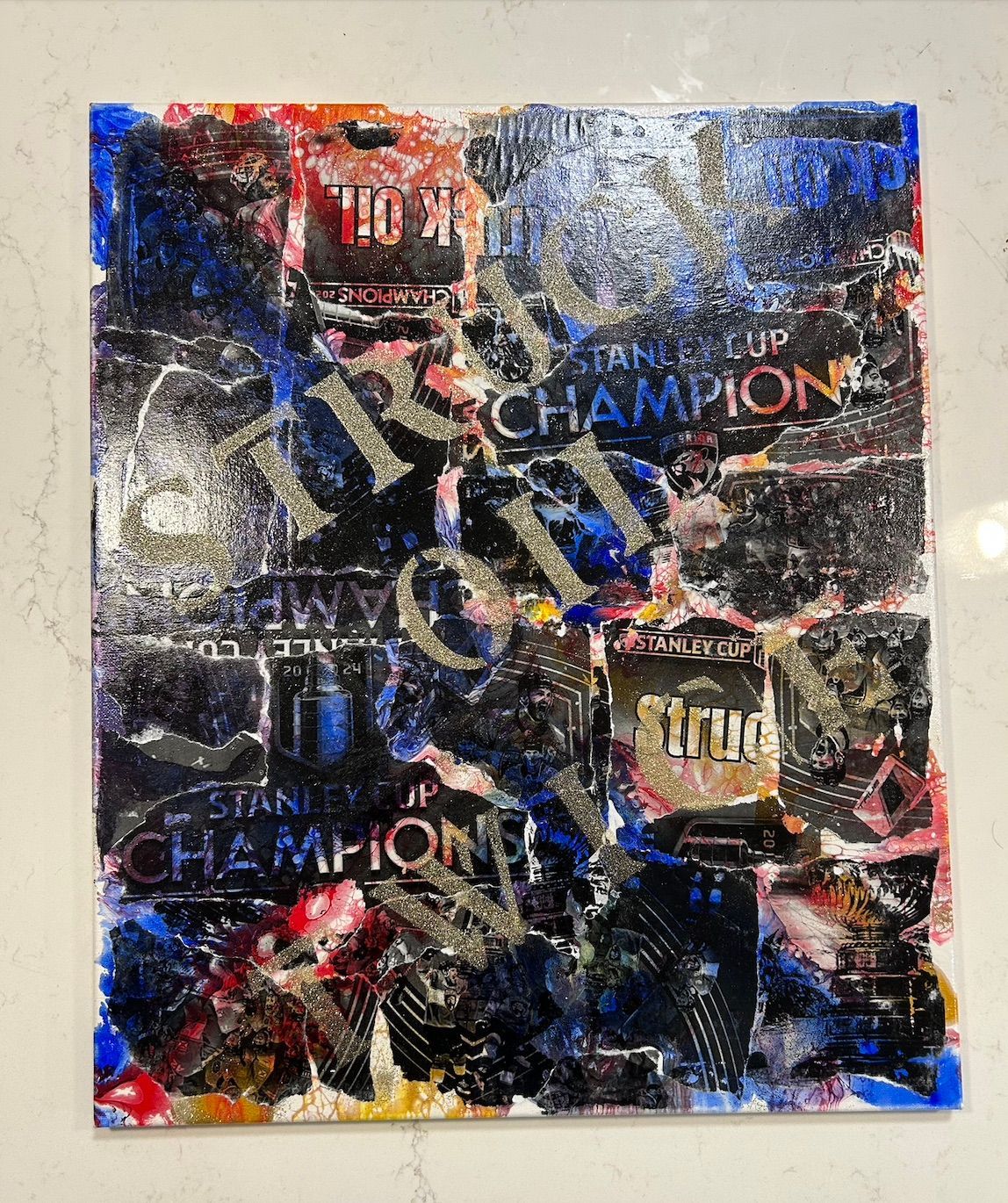 Abstract acrylic pour art with "CHAMPION" text in mixed media. Blue, black, silver, and red paint applied to a canvas.