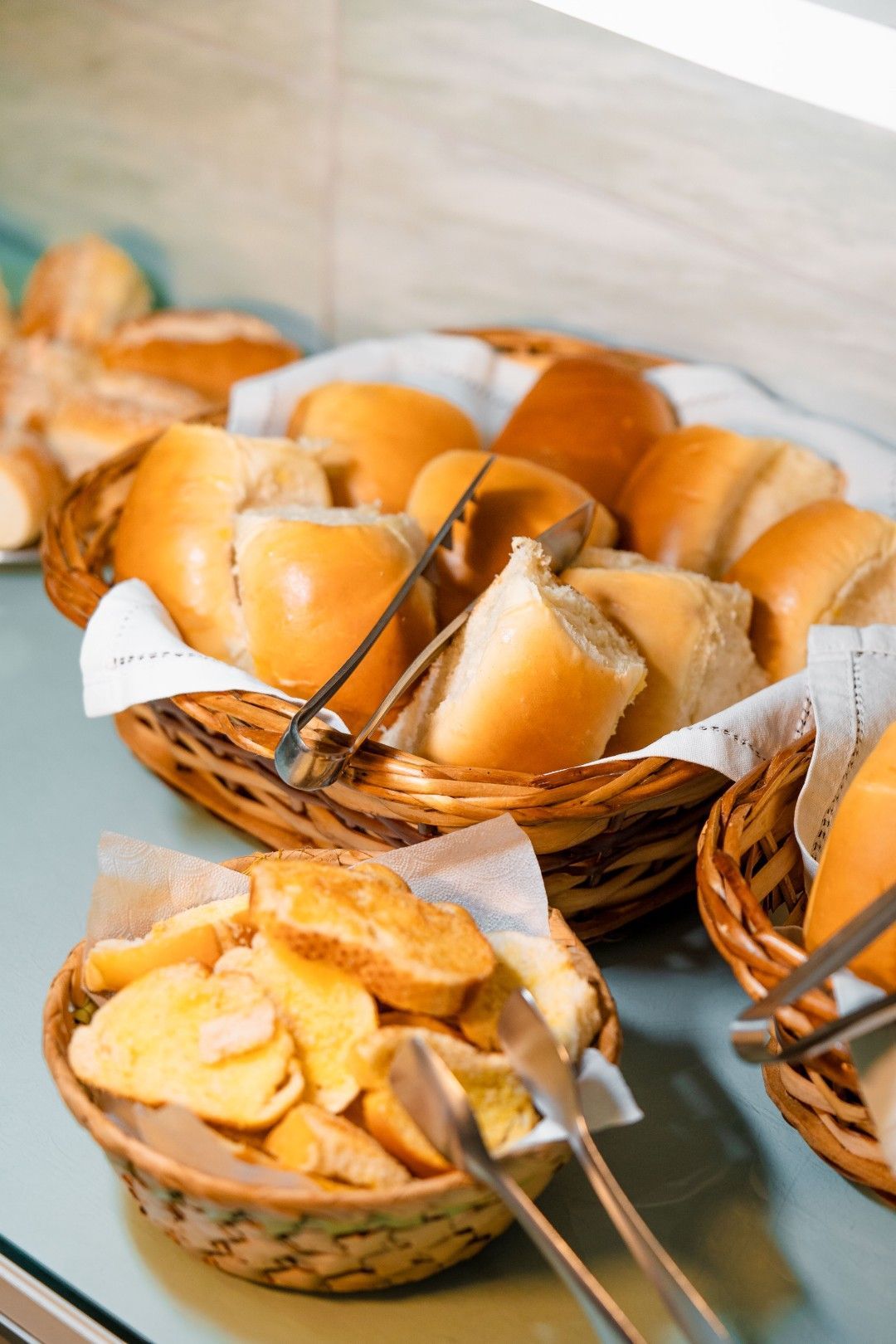 Cestas com diversos tipos de pães, incluindo pãezinhos e fatias torradas, acompanhadas de pinças para servir.