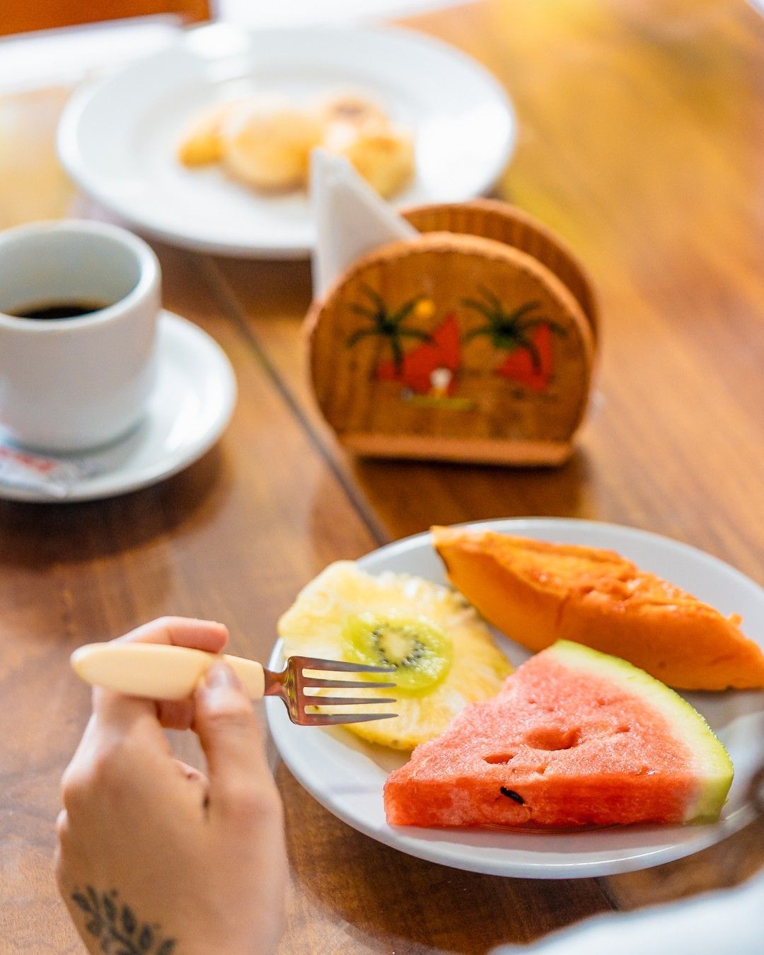 Prato de café da manhã com frutas, café e um doce. Mão segurando um garfo, mesa de madeira.
