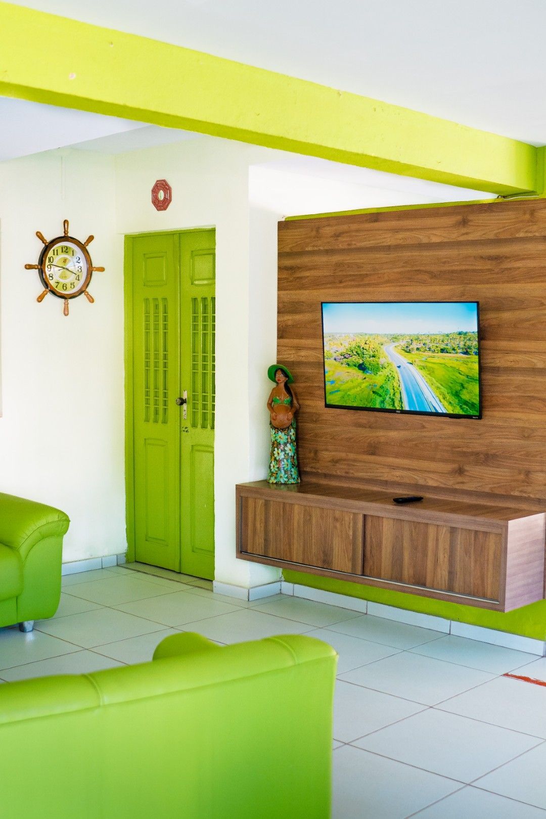 Sala de estar iluminada com sofá e porta verdes, parede com detalhes em madeira, TV e relógio náutico.