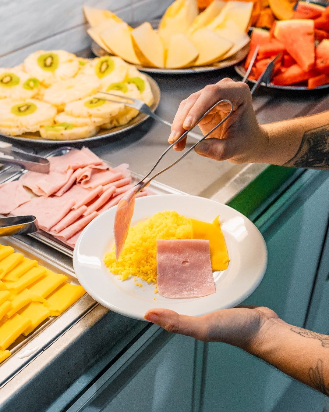 Pessoa usando pinças para servir presunto e outros alimentos de café da manhã em um bufê.