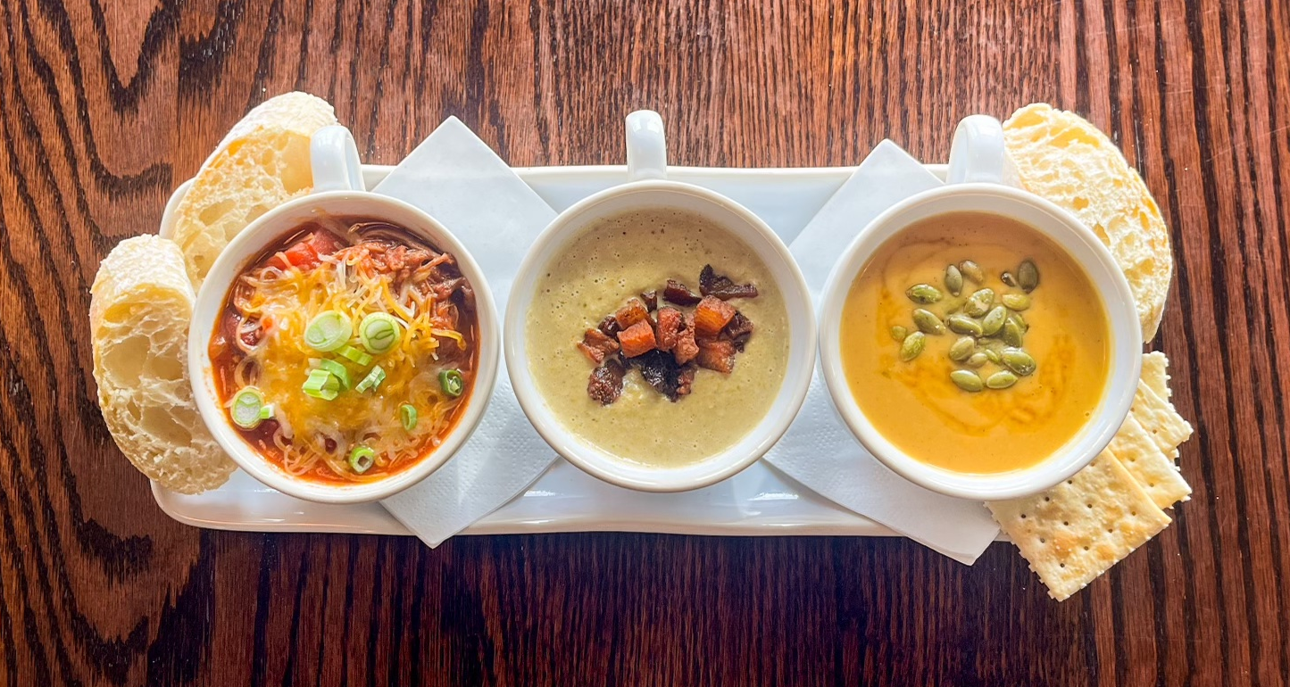 Three bowls of soup and crackers on a tray on a wooden table.
