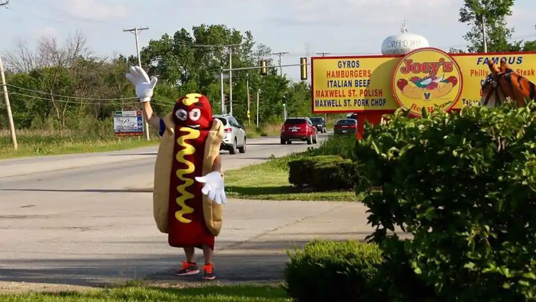 Person in hot dog costume waving by a road, Joey's restaurant sign in background.