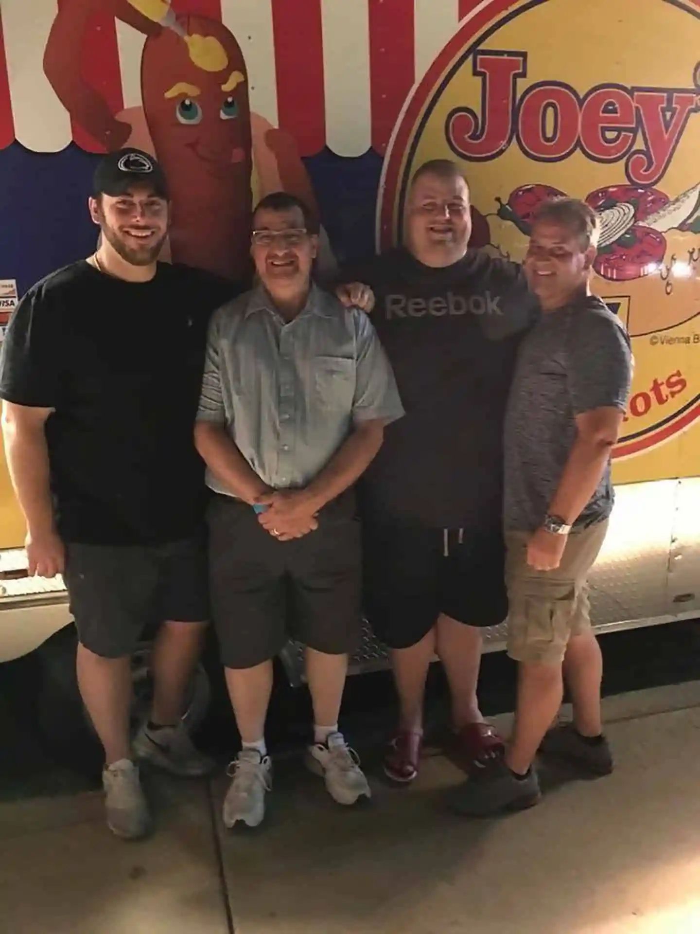 Four men pose in front of a hot dog stand with a cartoon hot dog on it.
